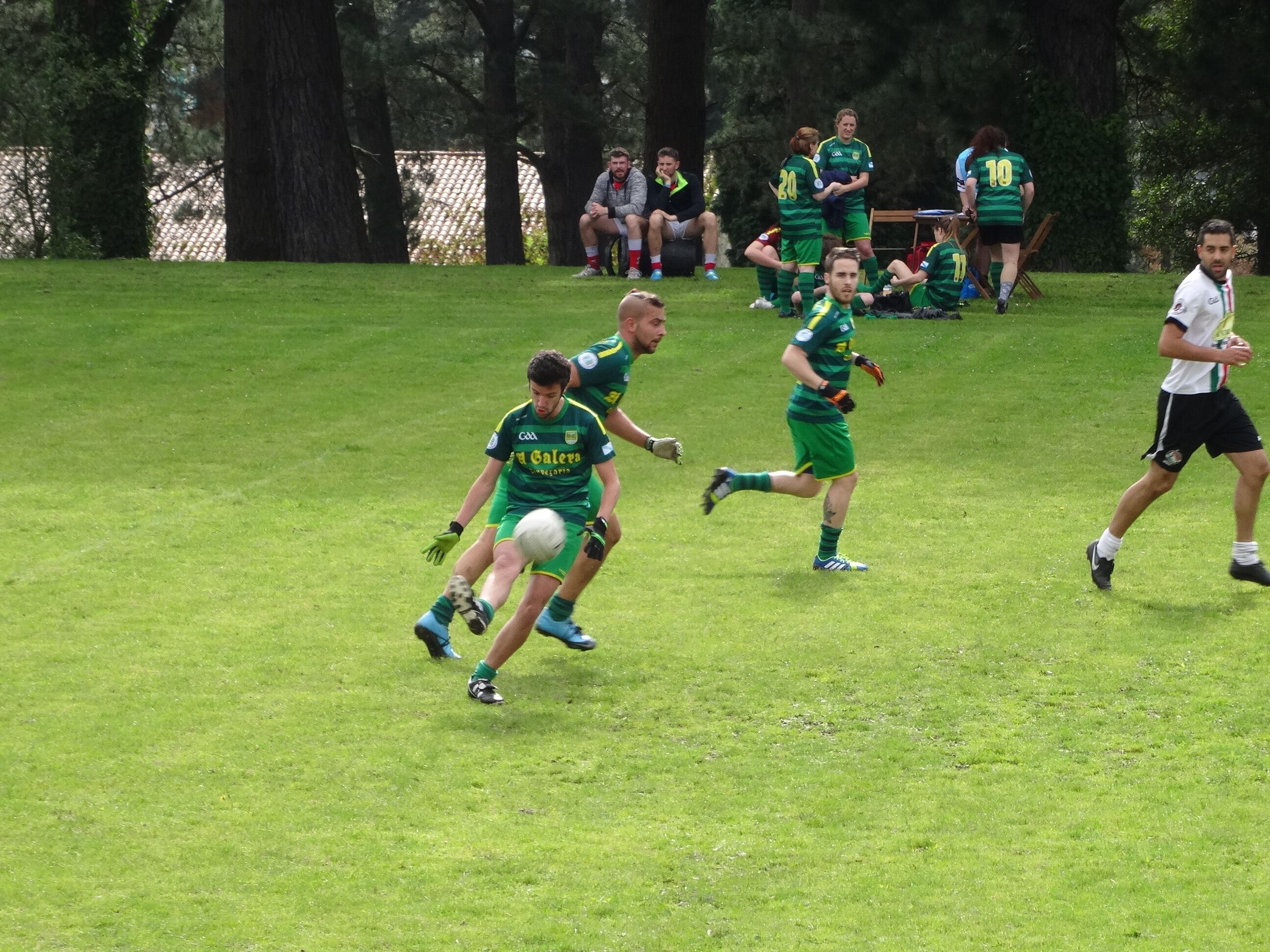 A grassy field with players in green and white uniforms, some sitting and others standing, likely during a sports event.