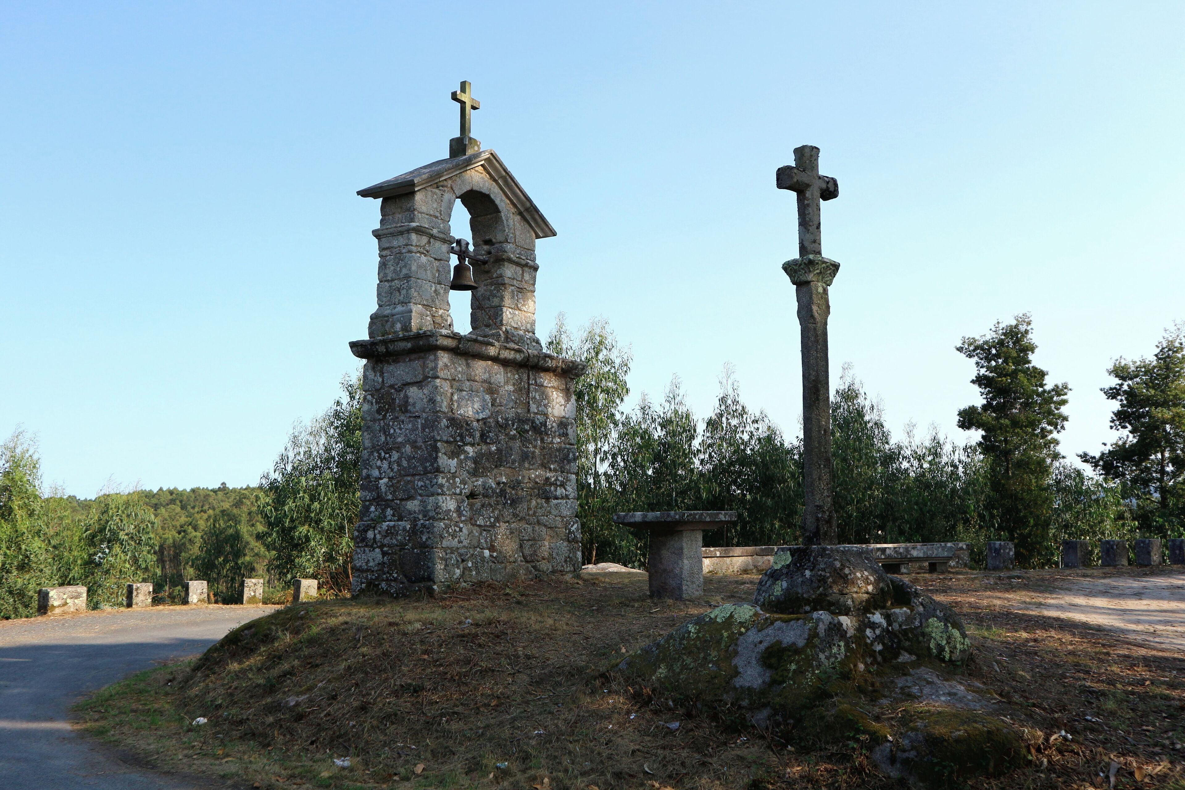 A stone bell tower with a cross on top, a large stone cross, and a stone bench in a wooded area.