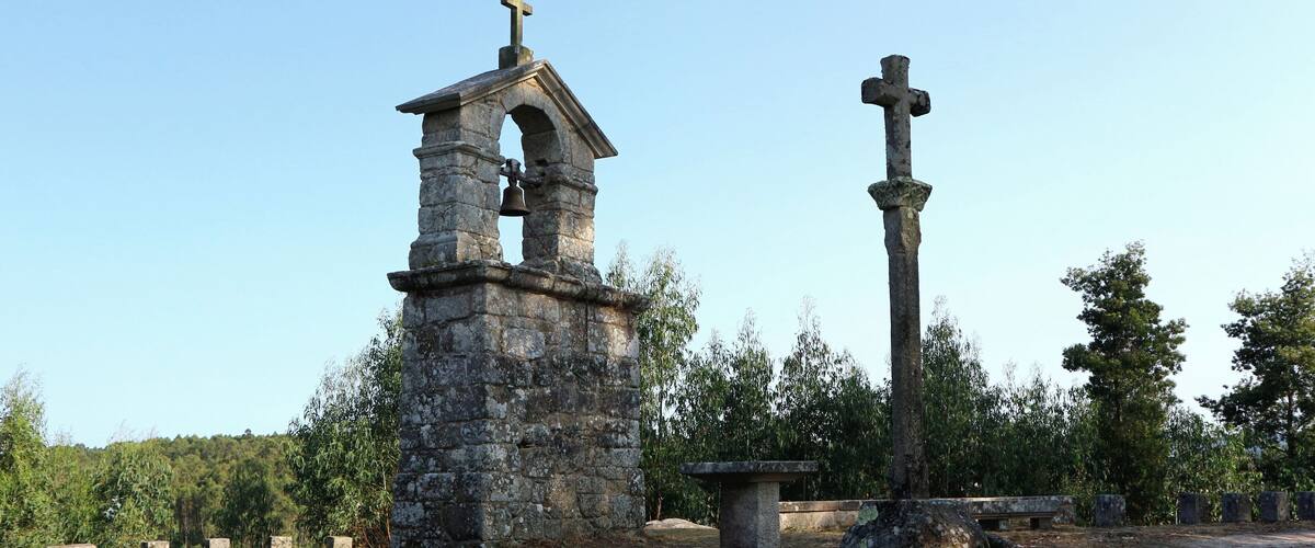 A stone bell tower with a cross on top, a large stone cross, and a stone bench in a wooded area.