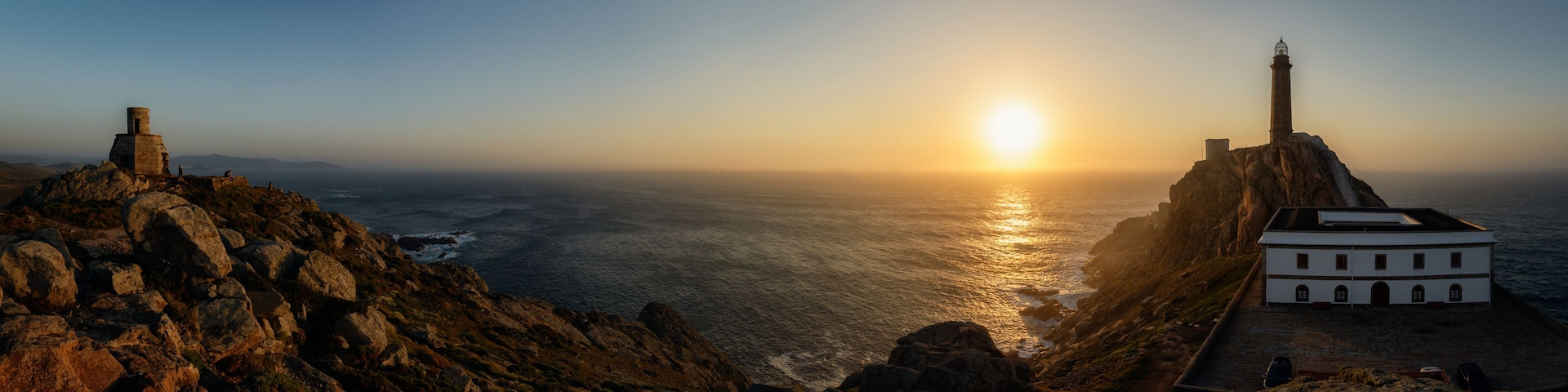 Un paisaje costero con un faro, terreno rocoso y un edificio blanco cerca del mar durante el atardecer.