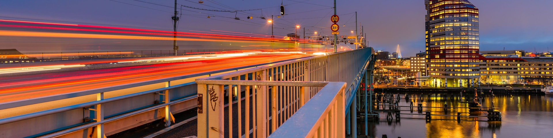 Uma ponte com rastros de luz de veículos em movimento, um corpo d'água refletindo luzes e um edifício moderno com janelas iluminadas.