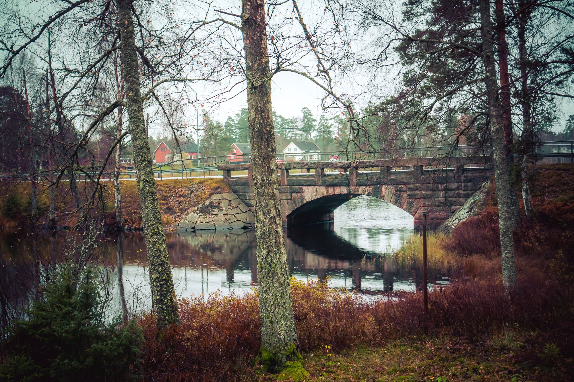 A stone bridge over a calm river, surrounded by trees and autumn foliage.