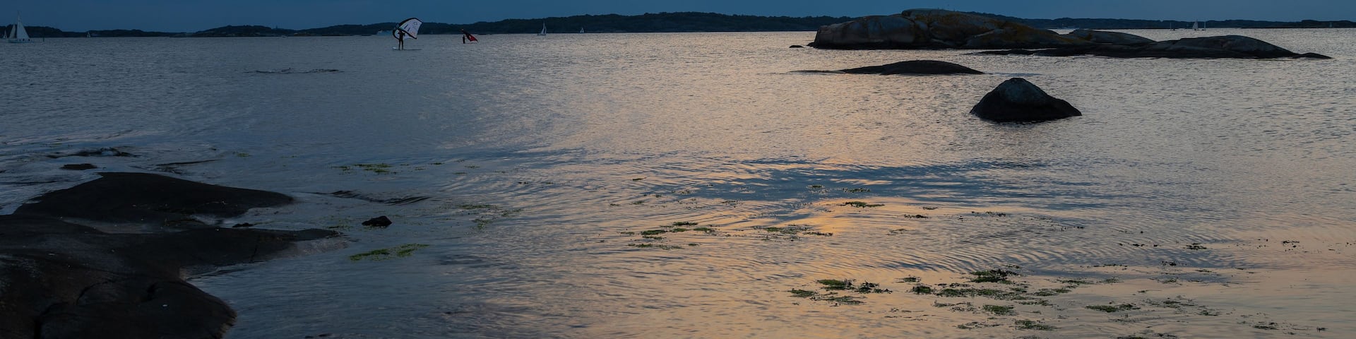 A calm sea with rocks and a sailboat in the distance under a cloudy sky.