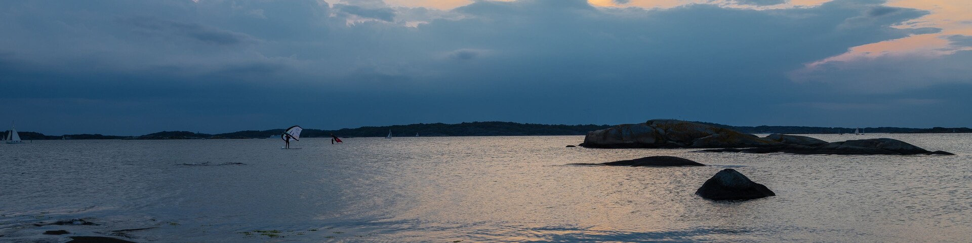 Mar en calma con rocas y un velero a lo lejos bajo un cielo nublado.