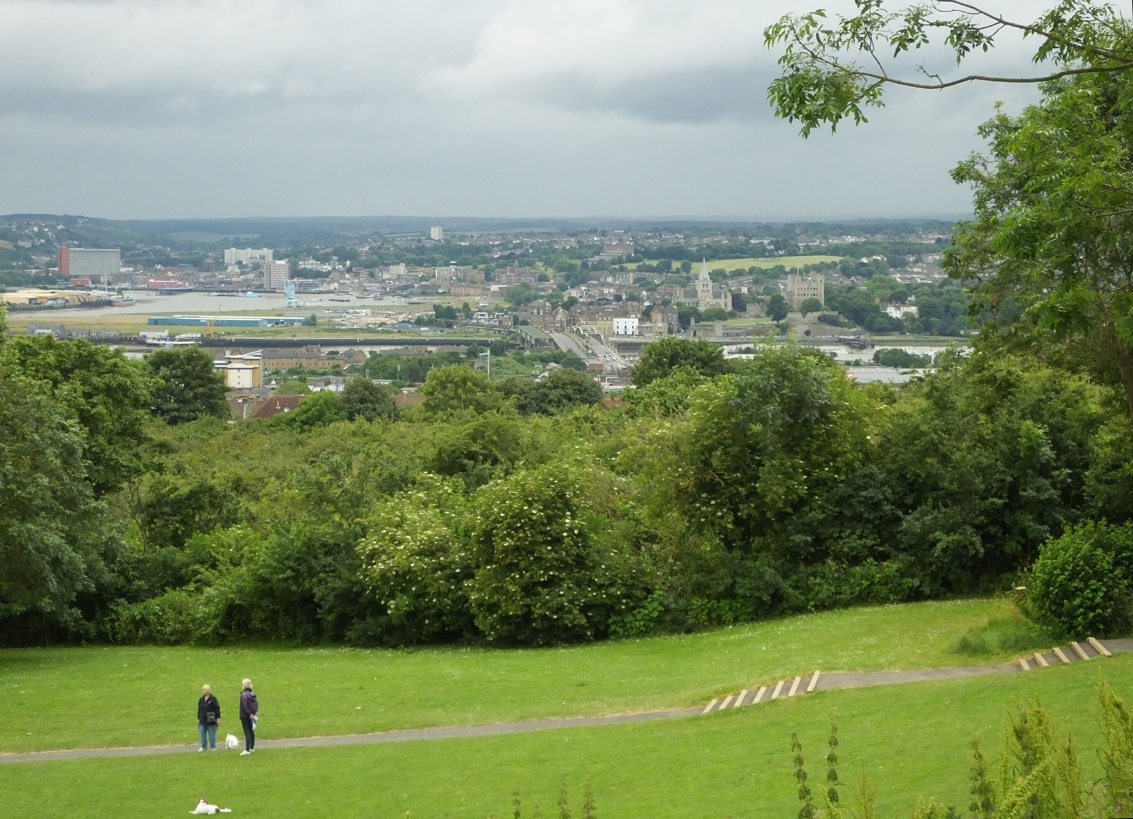 A park with two people walking, a river, and a cityscape in the background.