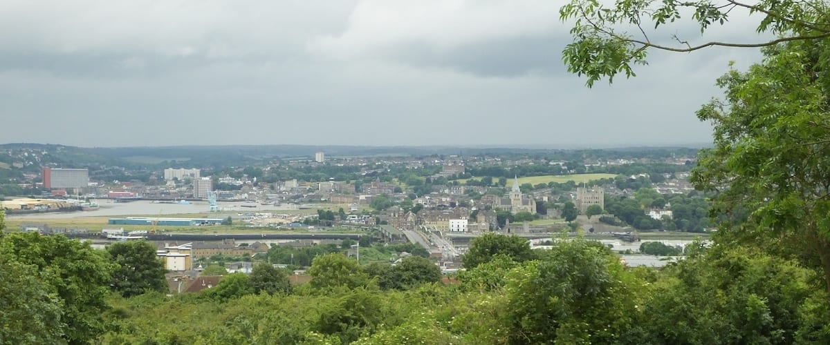 A park with two people walking, a river, and a cityscape in the background.