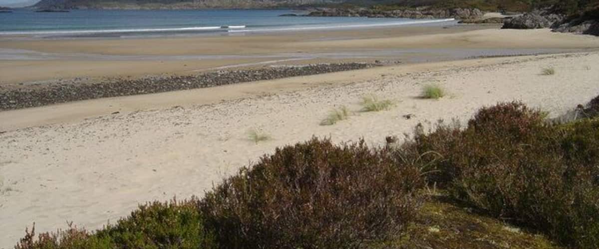 A coastal landscape with a sandy beach, rocky outcrops, and distant hills under a partly cloudy sky.