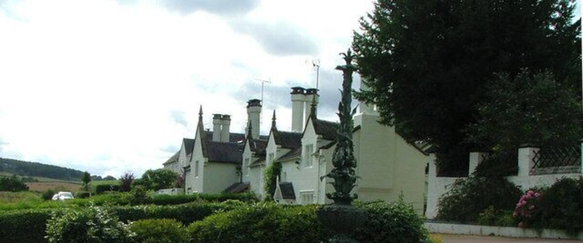 A white building with multiple chimneys, surrounded by well-maintained hedges and a circular pond with a fountain.