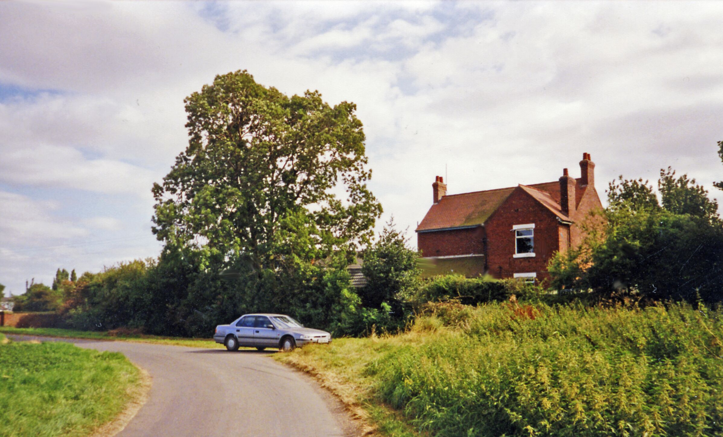 A rural road with a silver car, a large tree, and a brick house.