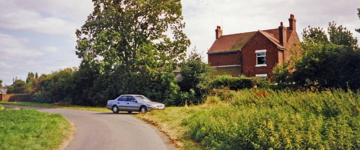 A rural road with a silver car, a large tree, and a brick house.