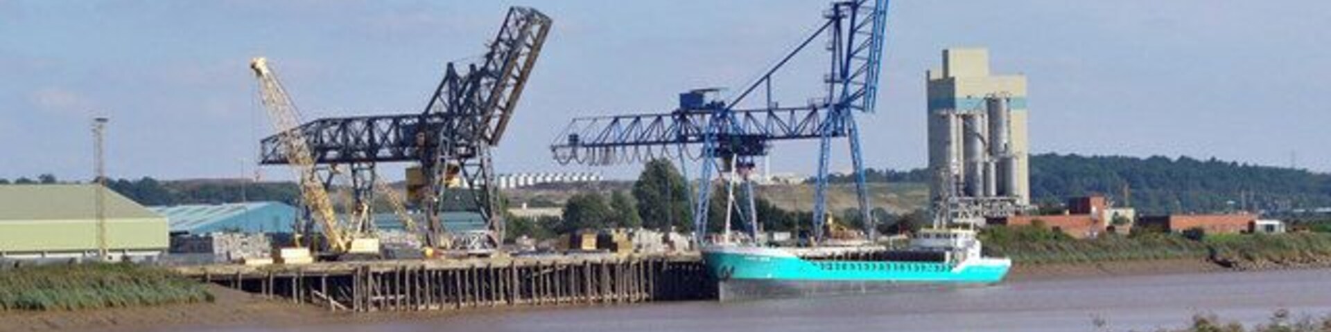 Um navio de carga atracado em um porto, com guindastes e edifícios industriais ao fundo.