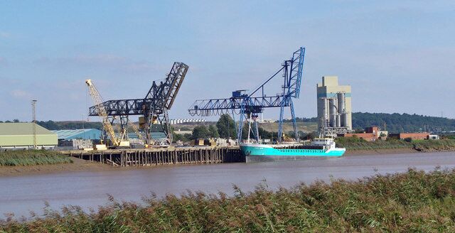 A cargo ship docked at a port with cranes and industrial buildings in the background.