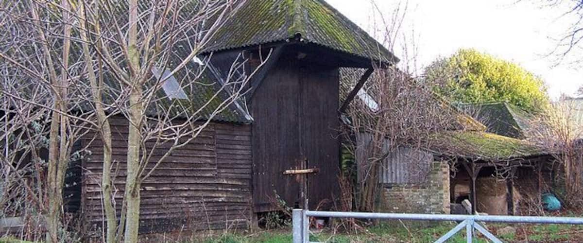 A rustic wooden barn with a metal gate in front, surrounded by bare trees and a small pond.