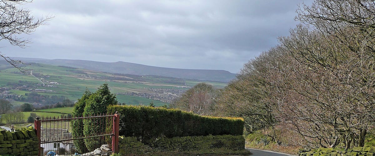 A rural road curving through a valley with stone walls and hedges on one side, and a view of distant fields and hills under a cloudy sky.