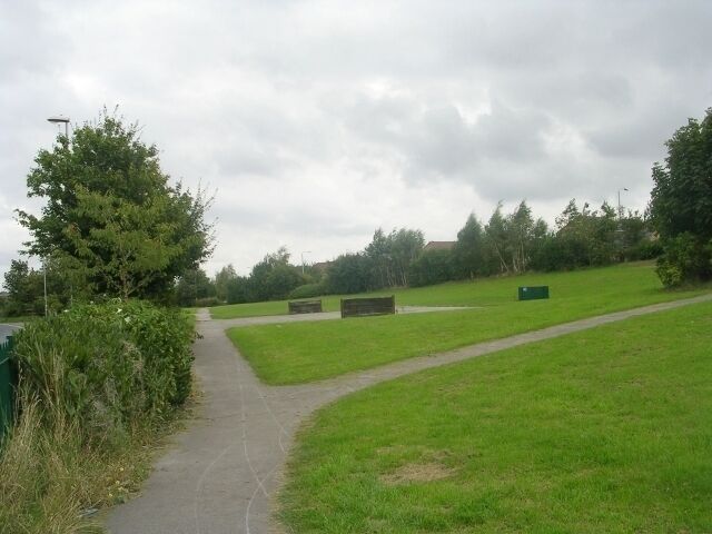 A paved path in a grassy park with trees and a cloudy sky.