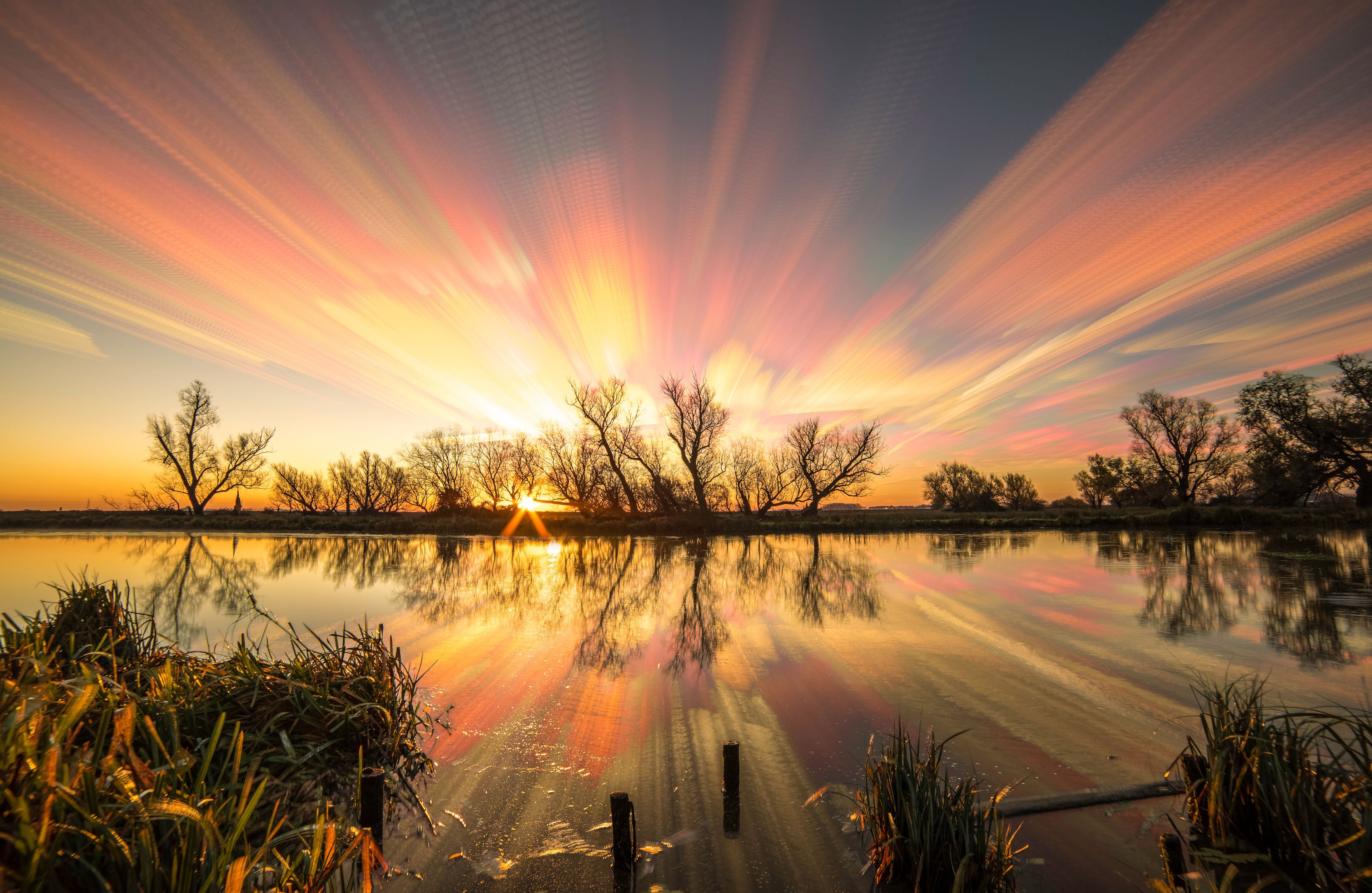 A sunset over a calm lake with reflections of trees and a dock.