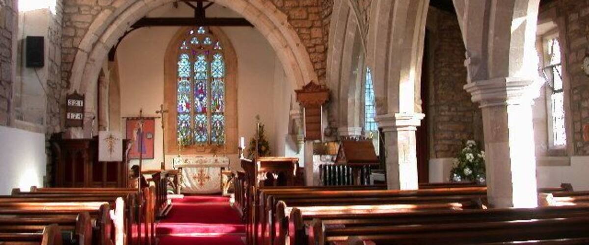 An interior of a church with stone walls, wooden pews, a red carpet, and a stained glass window.