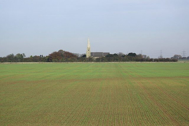 A rural landscape with a large open field, a church with a tall steeple, and a few buildings in the background.