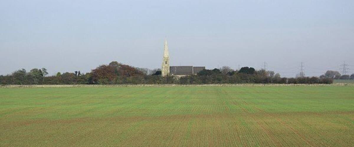 A rural landscape with a large open field, a church with a tall steeple, and a few buildings in the background.