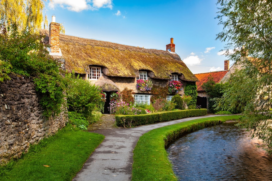 A quaint thatched-roof cottage with a stone wall, a narrow pathway, and a small water channel.