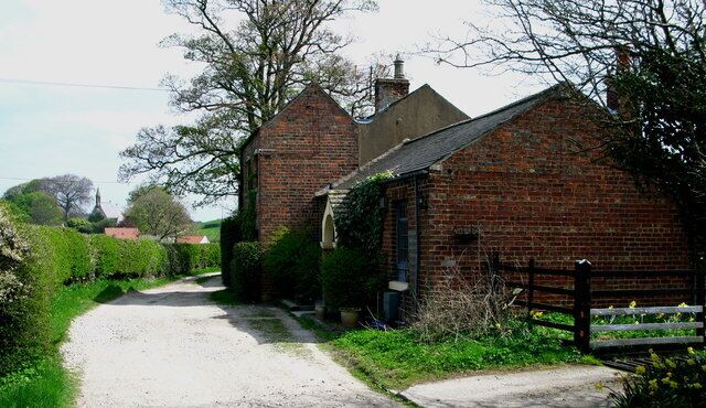 A rural road flanked by brick buildings, a wooden fence, and lush greenery.