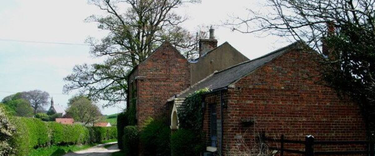 A rural road flanked by brick buildings, a wooden fence, and lush greenery.