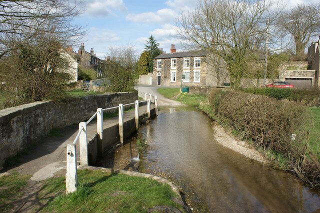 A narrow canal with a lock and a bridge, surrounded by greenery and buildings.