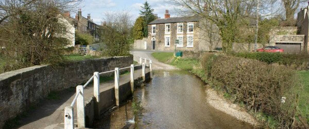 A narrow canal with a lock and a bridge, surrounded by greenery and buildings.