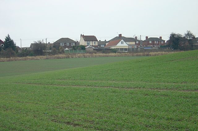A rural landscape with a green field, houses in the background, and a cloudy sky.