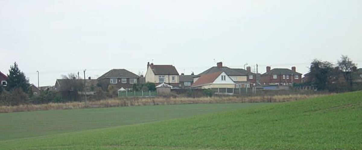 A rural landscape with a green field, houses in the background, and a cloudy sky.