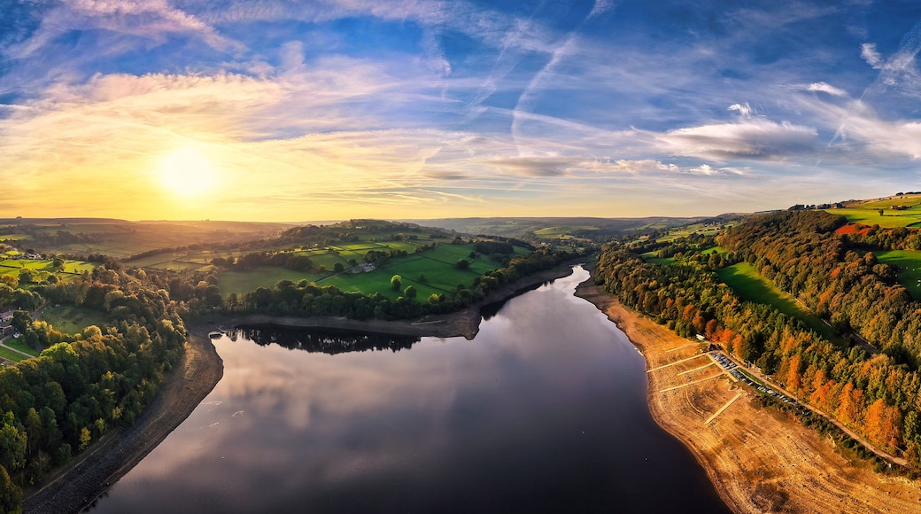 Ein Fluss schlängelt sich durch ein Tal mit Feldern und Bäumen, unter einem teils bewölkten Himmel.