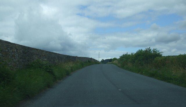 A narrow rural road flanked by stone walls and vegetation under a cloudy sky.