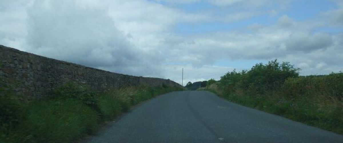 A narrow rural road flanked by stone walls and vegetation under a cloudy sky.