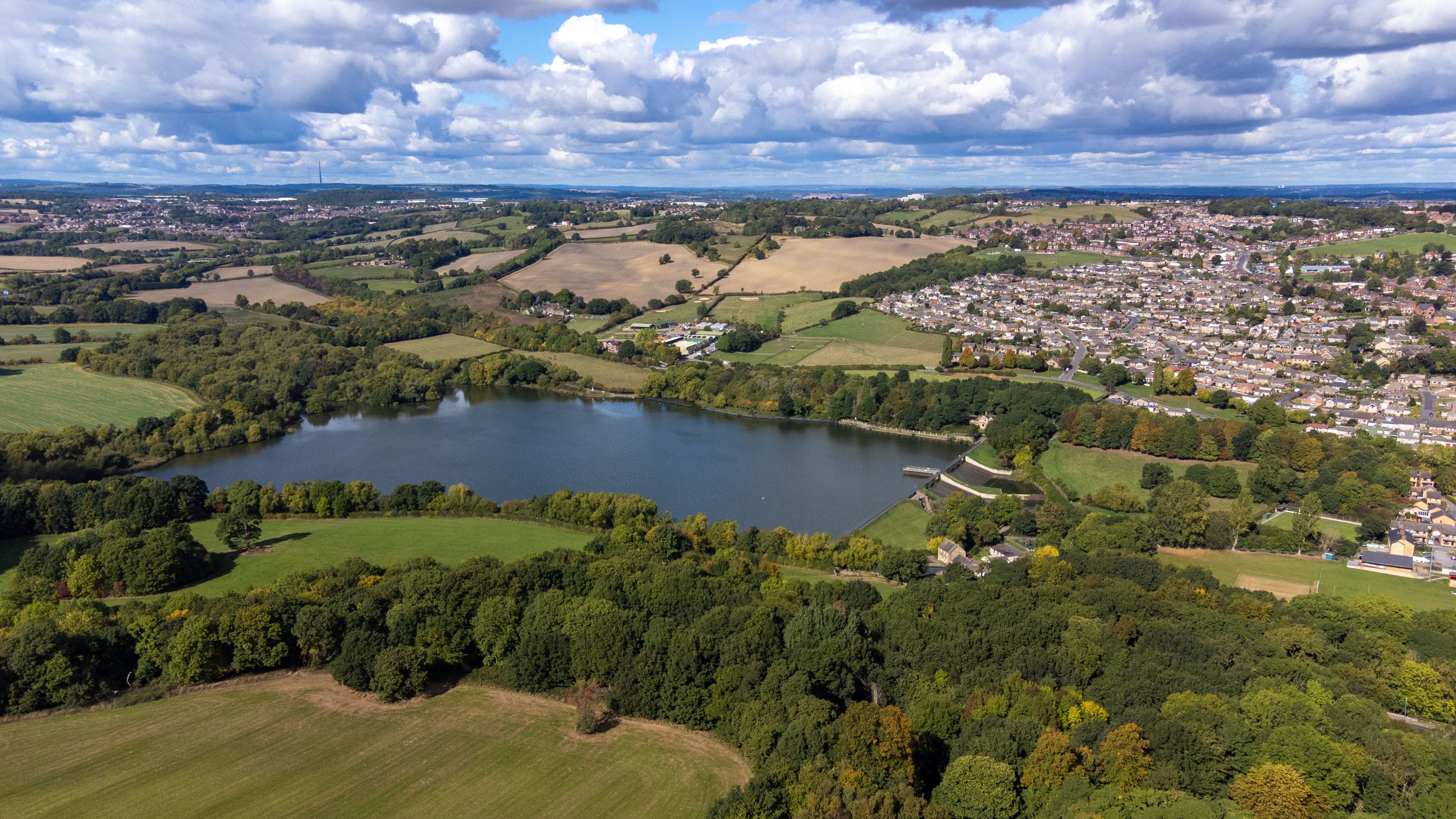 Ein See, umgeben von Grünflächen, eine Stadt mit Häusern und sanfte Hügel im Hintergrund.