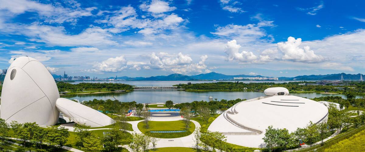 A modern architectural complex with unique white structures, a lake, and a city skyline in the background.