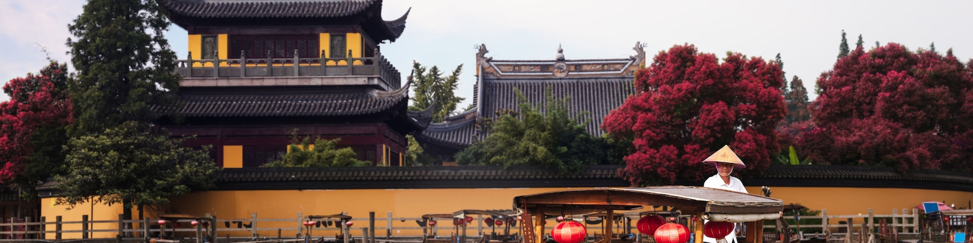 Un edificio tradicional chino con arquitectura ornamentada, un río con barcos y una persona con sombrero cónico.