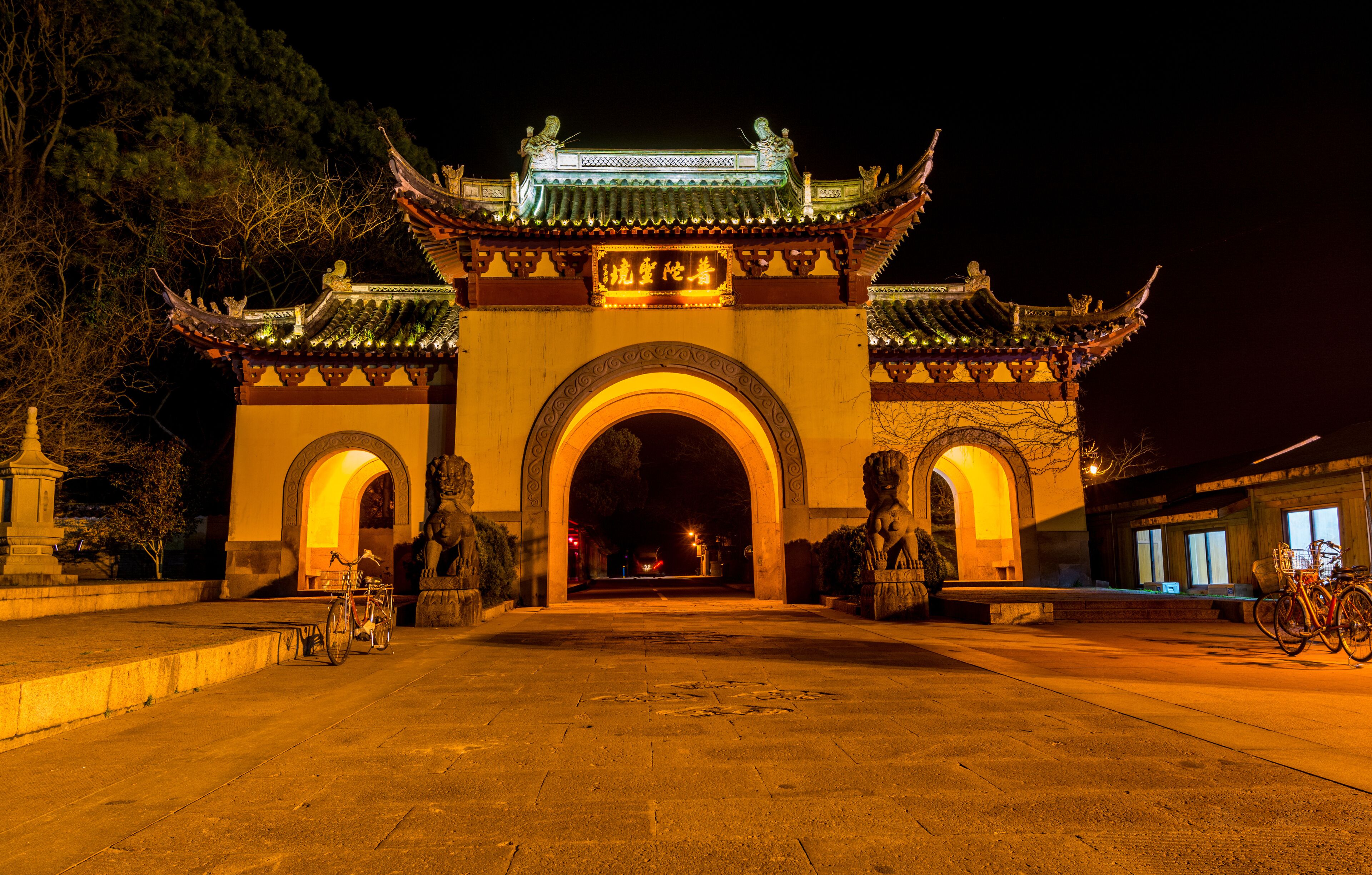 A traditional Chinese gate at night with intricate architecture and a bicycle parked nearby.