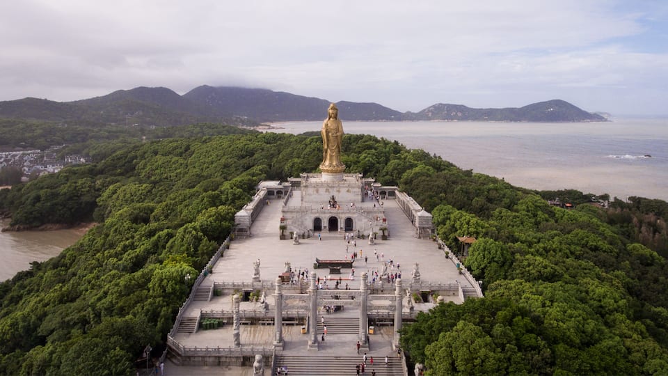 A large golden statue on a hilltop surrounded by lush greenery and overlooking a coastal area.