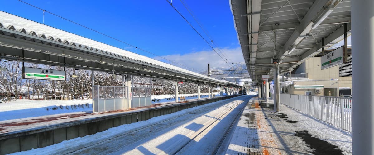 A snow-covered train station platform with tracks, overhead wires, and a clear blue sky.