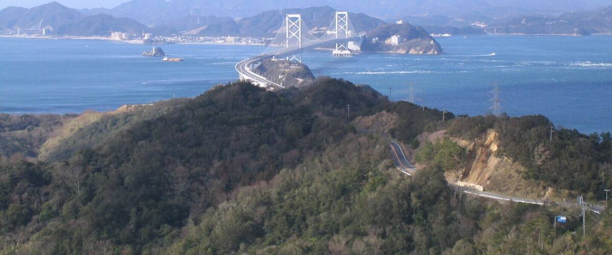 A coastal landscape with a prominent bridge, lush green hills, and distant mountains.