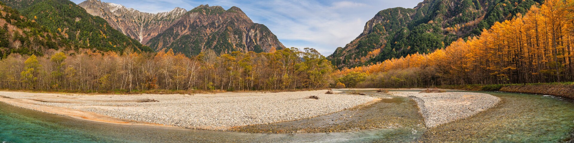 Um rio serpenteando por uma paisagem montanhosa com árvores em tons de outono e terreno rochoso.