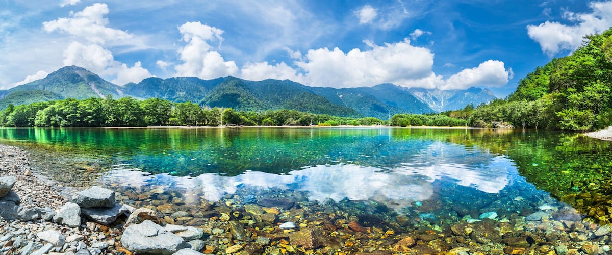A clear lake with a rocky shore, surrounded by lush green mountains and a partly cloudy sky.