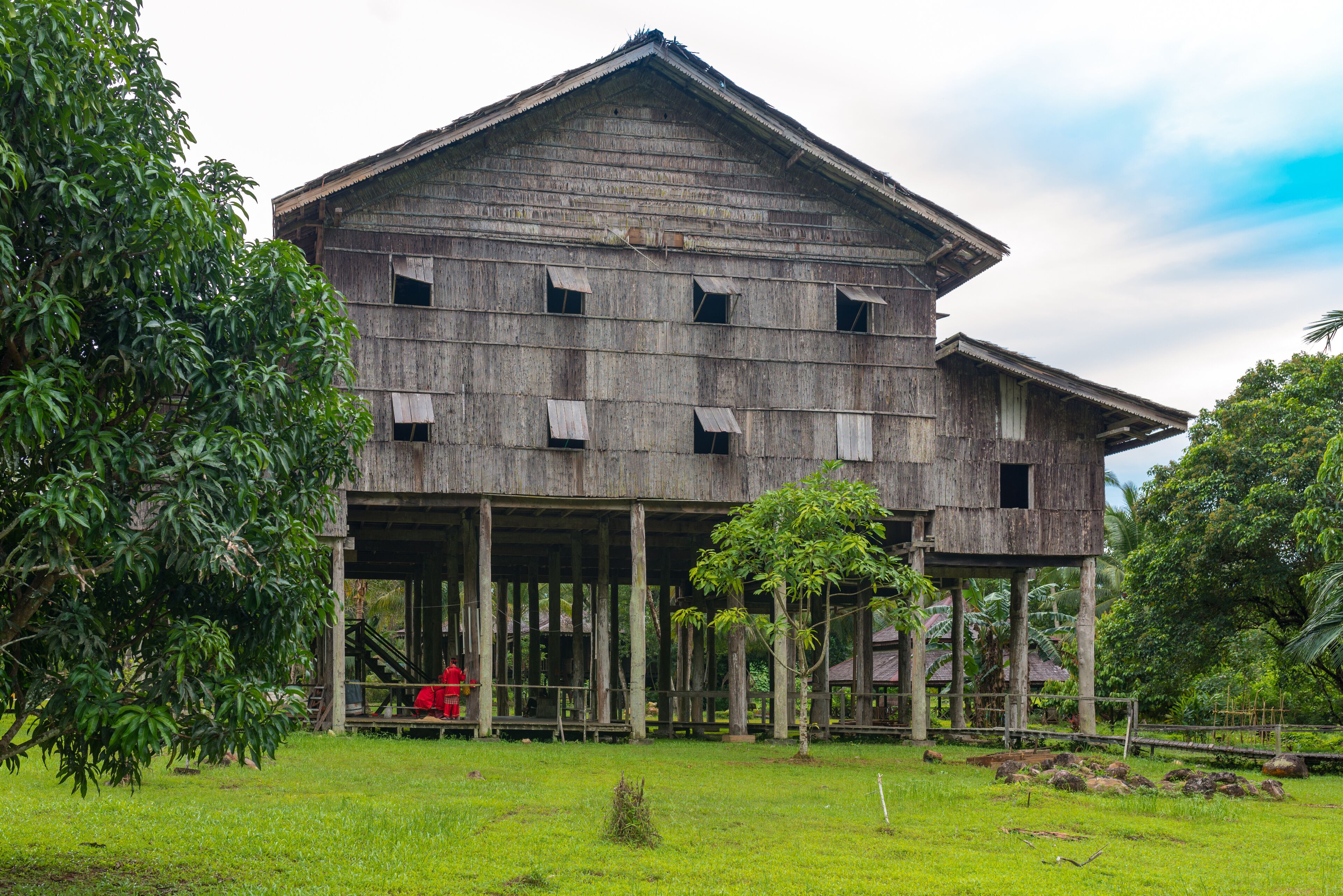 A large wooden building with multiple windows and a covered porch, surrounded by lush greenery and trees.