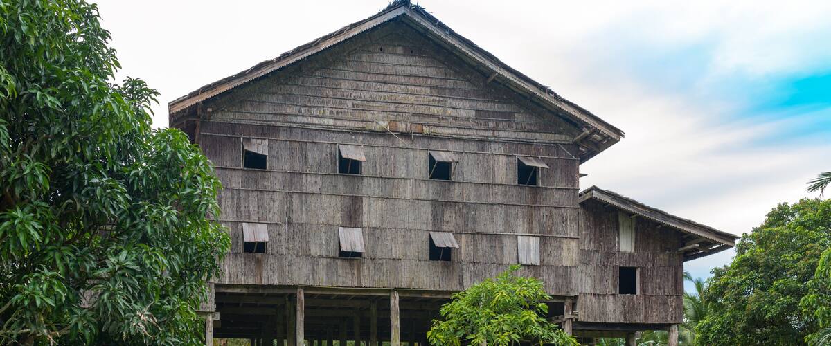 A large wooden building with multiple windows and a covered porch, surrounded by lush greenery and trees.
