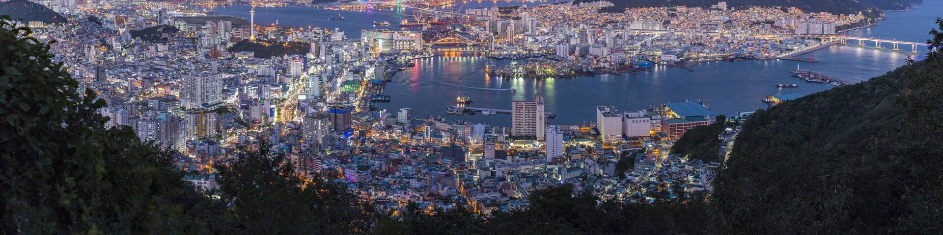 A panoramic view of a coastal city at dusk with illuminated buildings and a prominent bridge.
