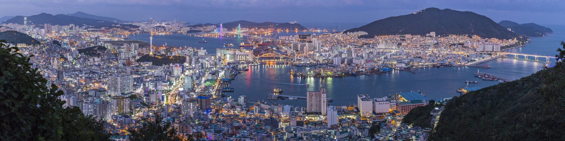 Vista panorâmica de uma cidade costeira ao anoitecer, com edifícios iluminados e uma ponte destacada.