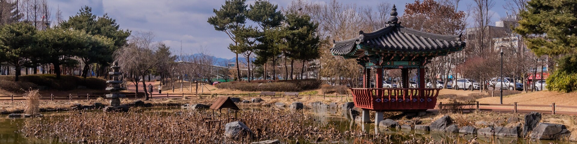 A traditional pavilion by a pond with reeds, trees, and a clear sky.