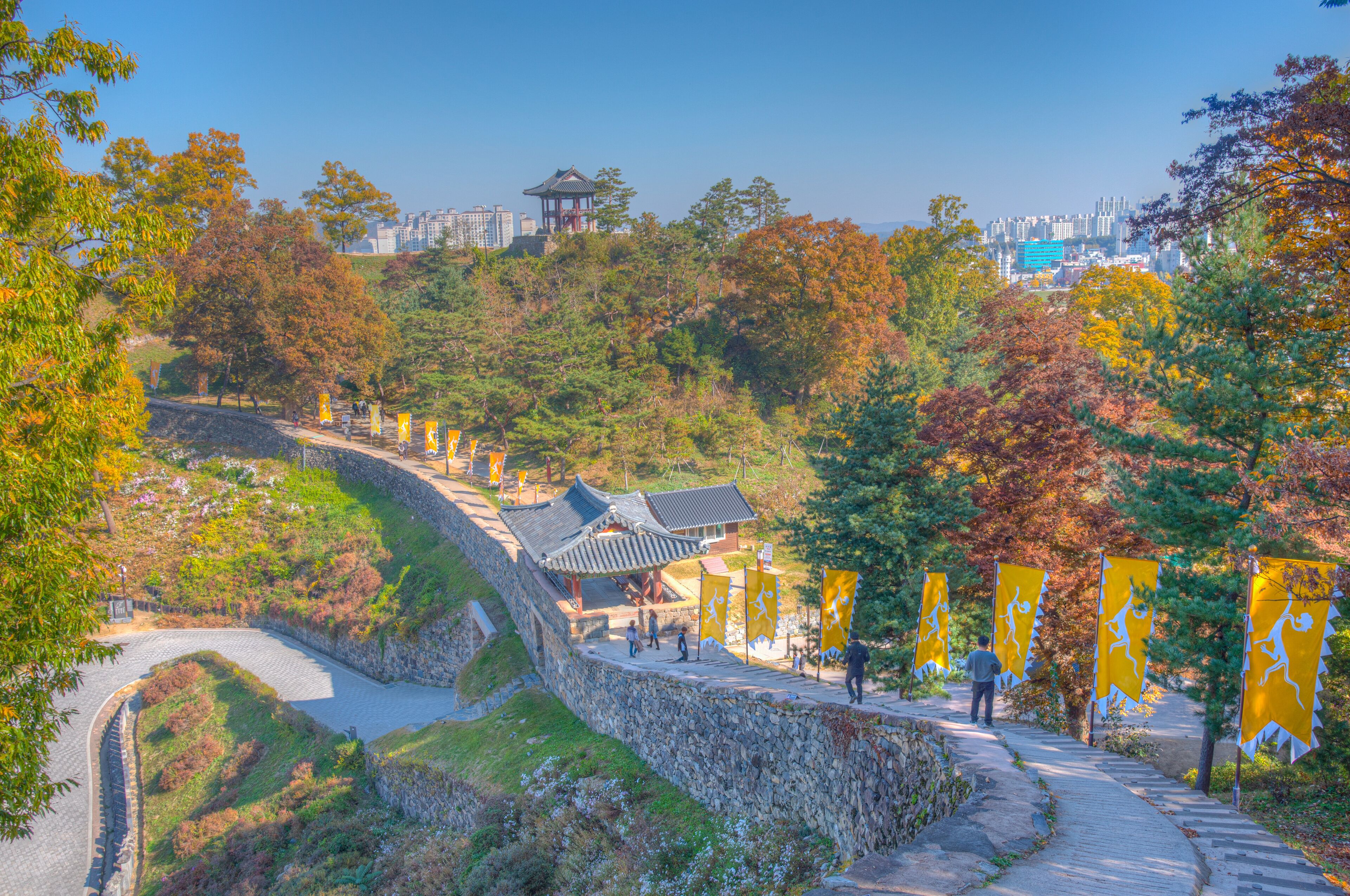 A traditional Korean pavilion, stone walls, and a pathway with yellow flags.