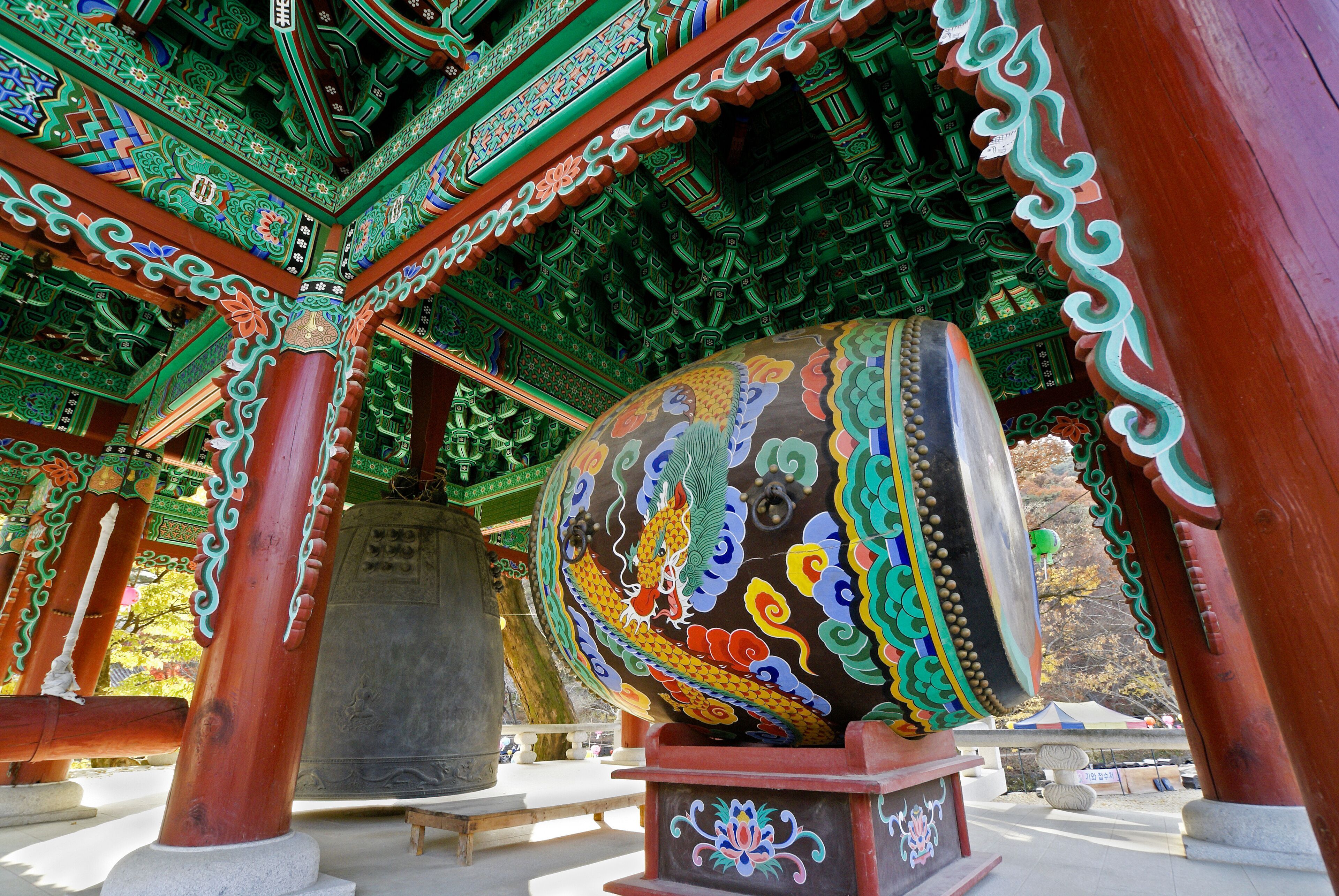 A traditional temple with ornate green and red architecture, a large bell, and a stone monument.