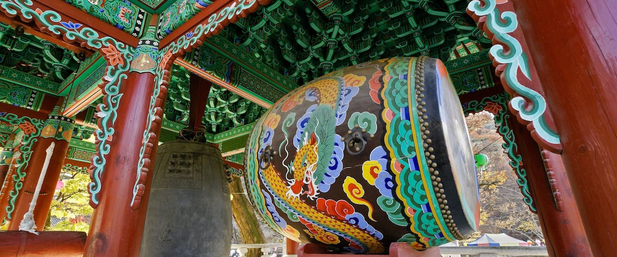 A traditional temple with ornate green and red architecture, a large bell, and a stone monument.